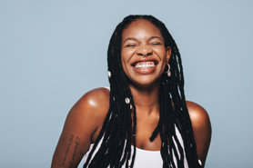 A woman with long, black, beaded dreadlocks and a septum ring smiles broadly against a light blue background. She wears a white tank top and hoop earrings, showing a tattoo on her left arm.