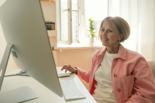 Smiling middle aged woman sitting at a desk in a bright home office using a desktop computer while studying or retraining online.