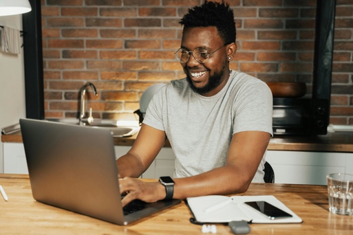 Man Smiling While Working On A Laptop From A Kitchen Table At Home