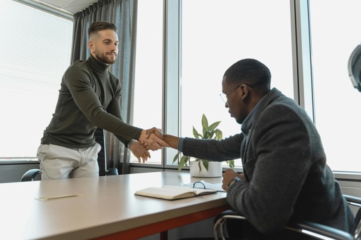 A graduate job candidate shakes hands with an interviewer across a desk in a bright office.