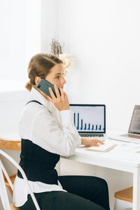 A person sitting at a desk taking a phone call. On the desk is two laptops and some papers.