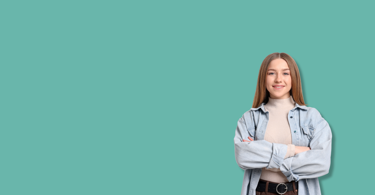 A young woman with long hair stands smiling with arms crossed, wearing a light denim shirt and turtleneck, against a plain teal background with ample empty space to her left.