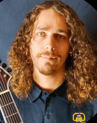 A man with long curly hair and a goatee stands against a dark background, holding an electric guitar. He wears a navy blue collared shirt with a yellow and white logo on the chest.