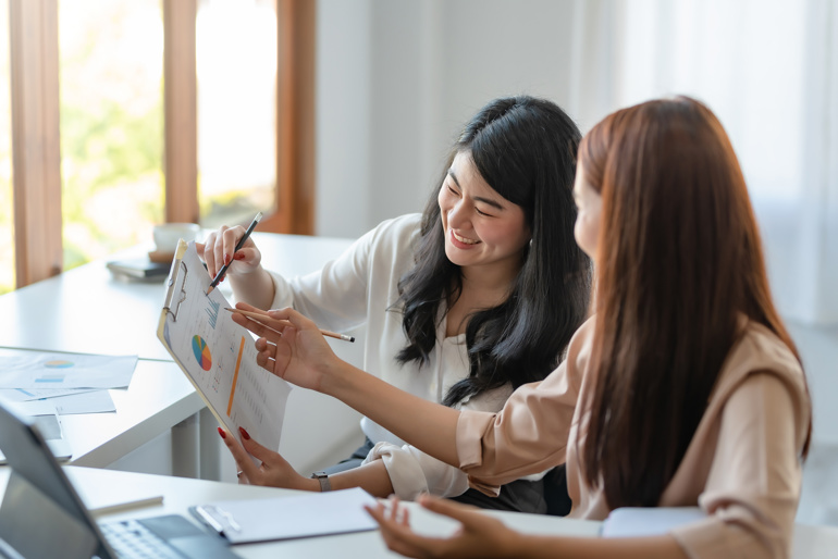 Two women sitting together looking at a clipboard, on which there's a document with project management statistic infographics on it, One is mentoring the other as a project management apprentice.