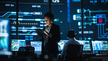 A woman stands using a tablet in a high-tech control room with large digital screens displaying data, while a seated man with headphones works at a computer in the background.