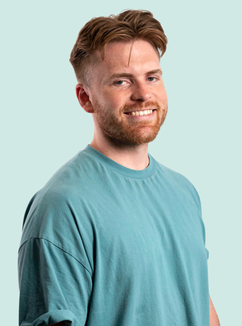 Smiling young man with short, light brown hair and a beard, wearing a teal t-shirt, standing against a plain light blue background.