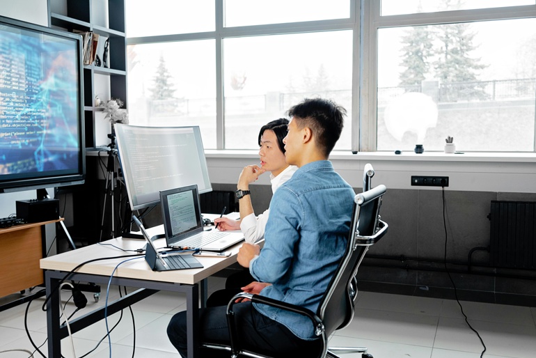Two young, male cyber professionals collaborating on some work on multiple computer and device screens.