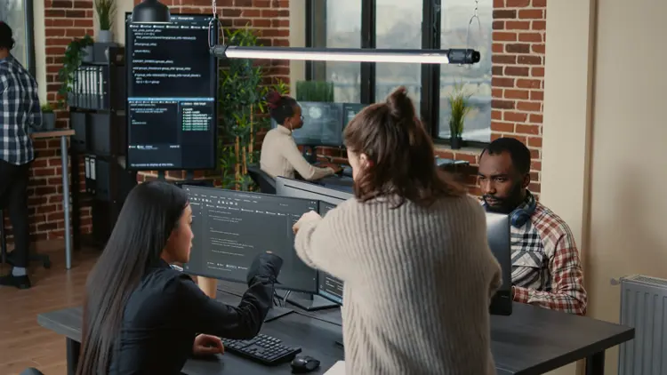 Four people work at computers in a modern office with brick walls. Two women discuss code on a monitor, while two others focus on their screens in the background. Multiple monitors display programming code.