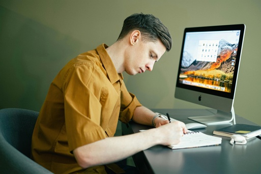 Person working at a desk using a computer, while taking notes in a notebook. 