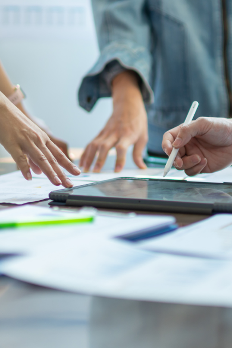 People's hands as they collaborate around a digital tablet device and paperwork, working on managing a project.