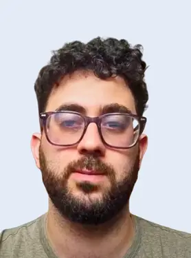 A man with short, curly dark hair, a beard, and glasses is looking at the camera against a plain light background. He is wearing a light grey t-shirt.