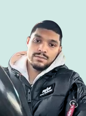 A man with short black hair and a trimmed beard looks at the camera. He wears a black puffer jacket over a gray hoodie, and the background is a plain light blue.