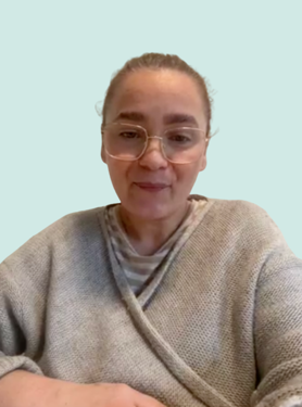 A person with light skin and glasses, wearing a grey sweater over a striped shirt, looks at the camera while sitting against a plain light blue background.