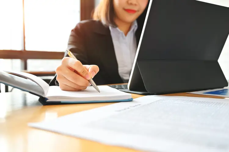 A woman in business attire writes in a notebook while working on a laptop at a desk covered with papers and documents, in a sunlit office setting.