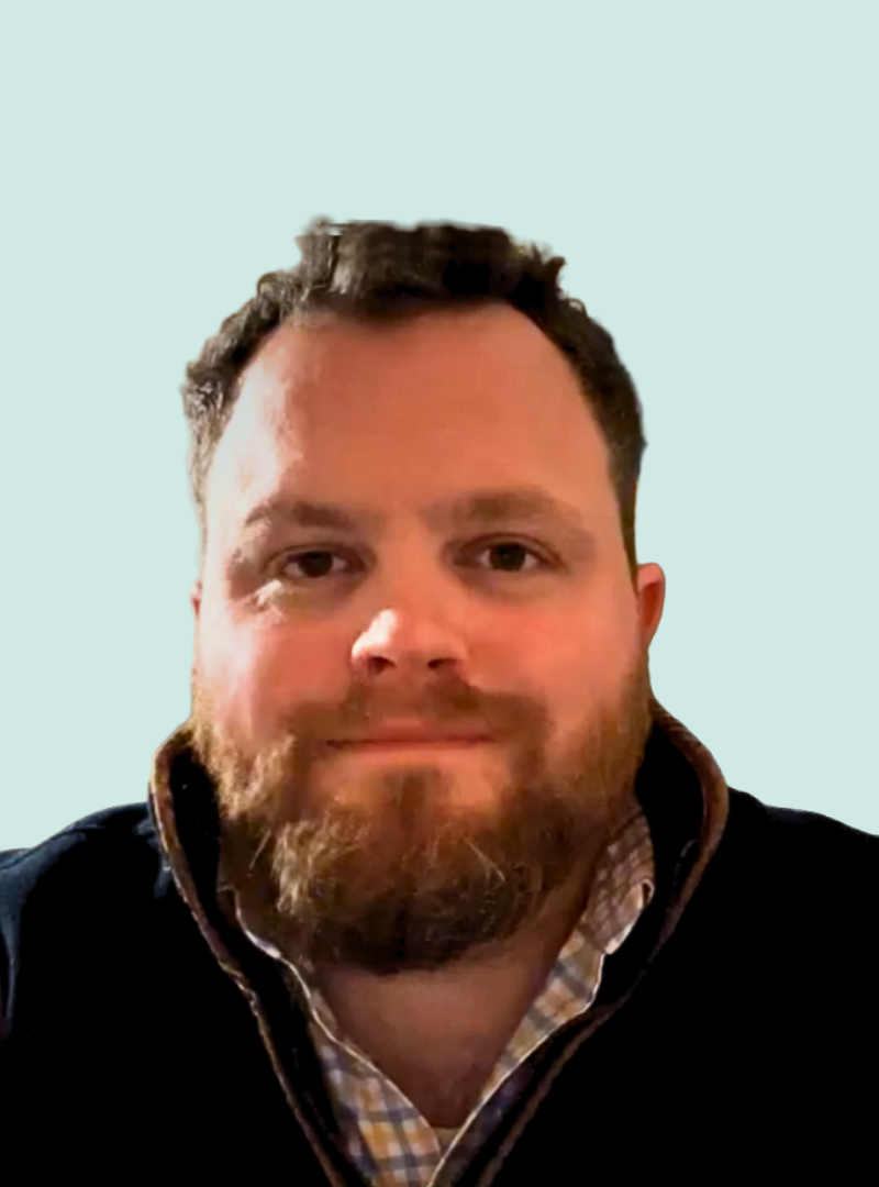 A bearded man with short brown hair, wearing a collared shirt and dark jacket, looks directly at the camera against a light blue background.