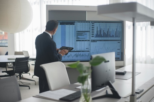 Professional reviewing data dashboards on a large office screen while holding a tablet.