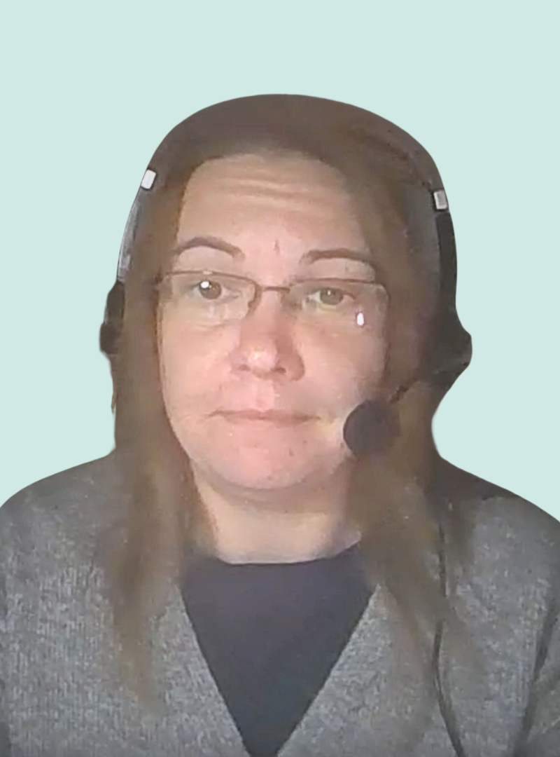 A person with long brown hair, wearing glasses and a headset with a microphone, looks at the camera. They are dressed in a dark top and gray sweater, against a light blue background.