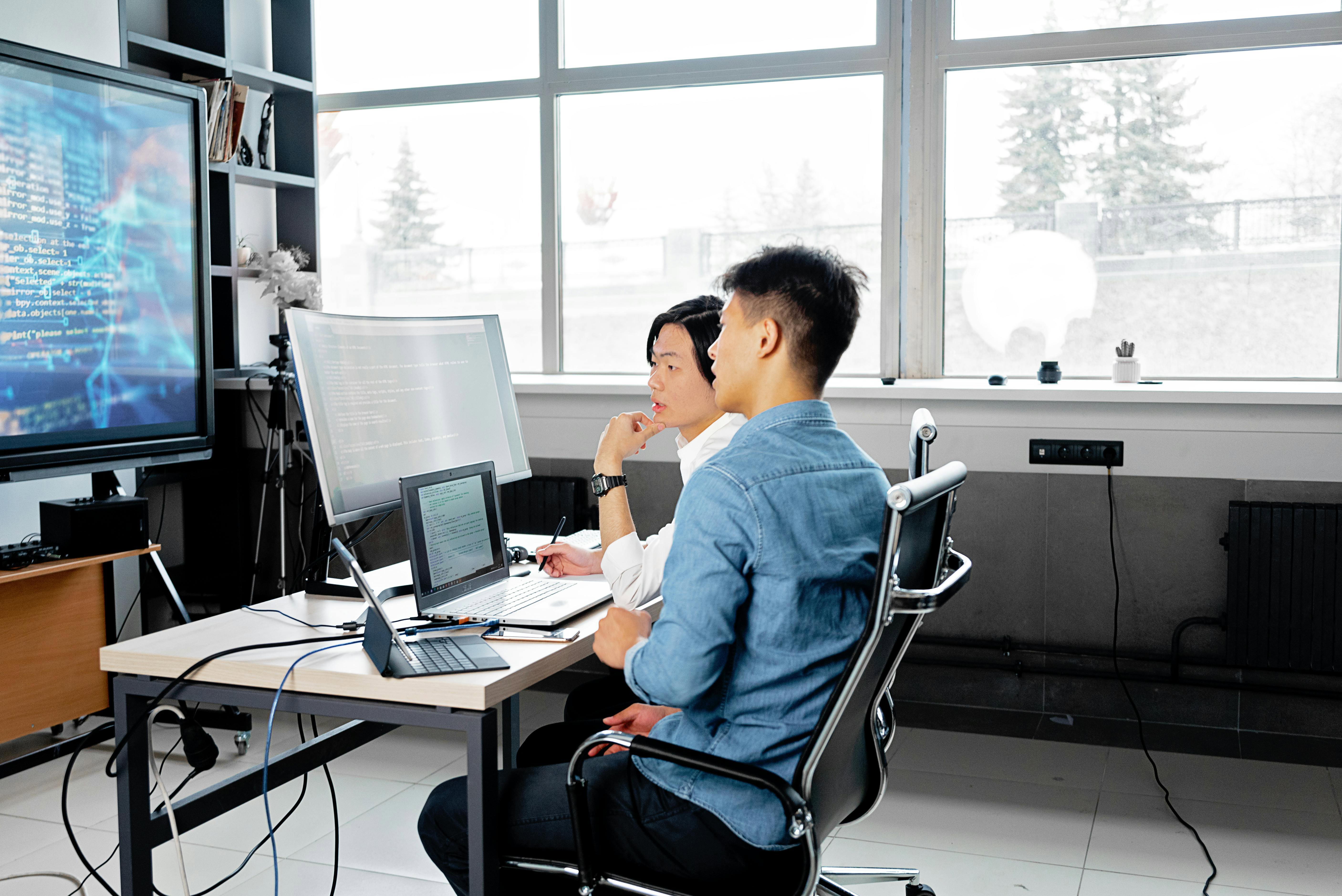 Two young, male cyber professionals collaborating on some work on multiple computer and device screens. 