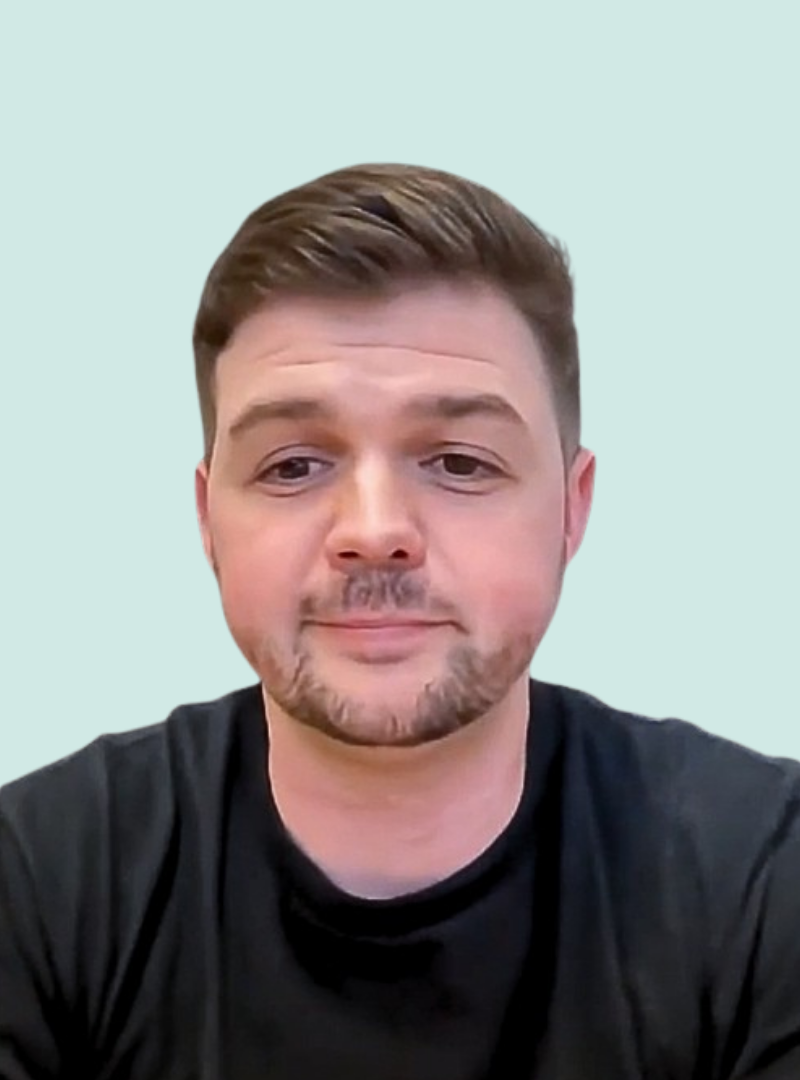 A man with short brown hair, trimmed beard, and mustache, wearing a black shirt, is facing the camera against a light blue background, with a neutral expression.