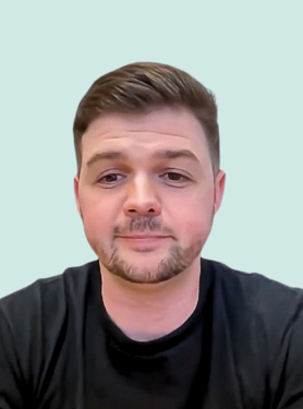 A man with short brown hair, trimmed beard, and mustache, wearing a black shirt, is facing the camera against a light blue background, with a neutral expression.