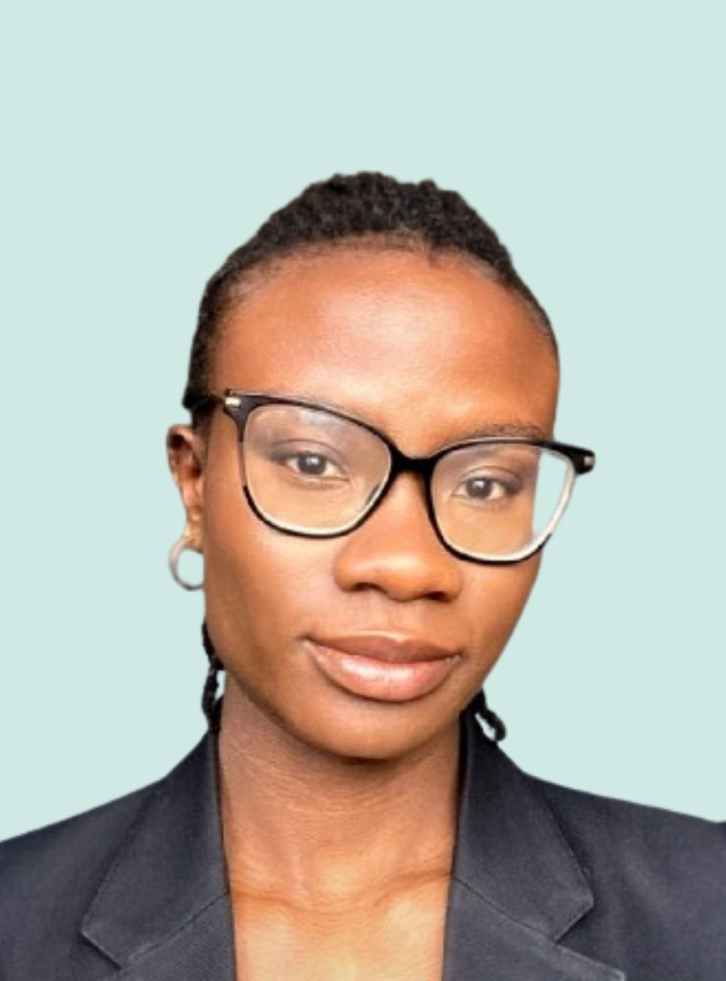 A person with glasses and short braids, wearing a dark blazer, looks at the camera with a neutral expression against a light blue background.