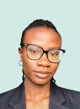 A person with glasses and short braids, wearing a dark blazer, looks at the camera with a neutral expression against a light blue background.